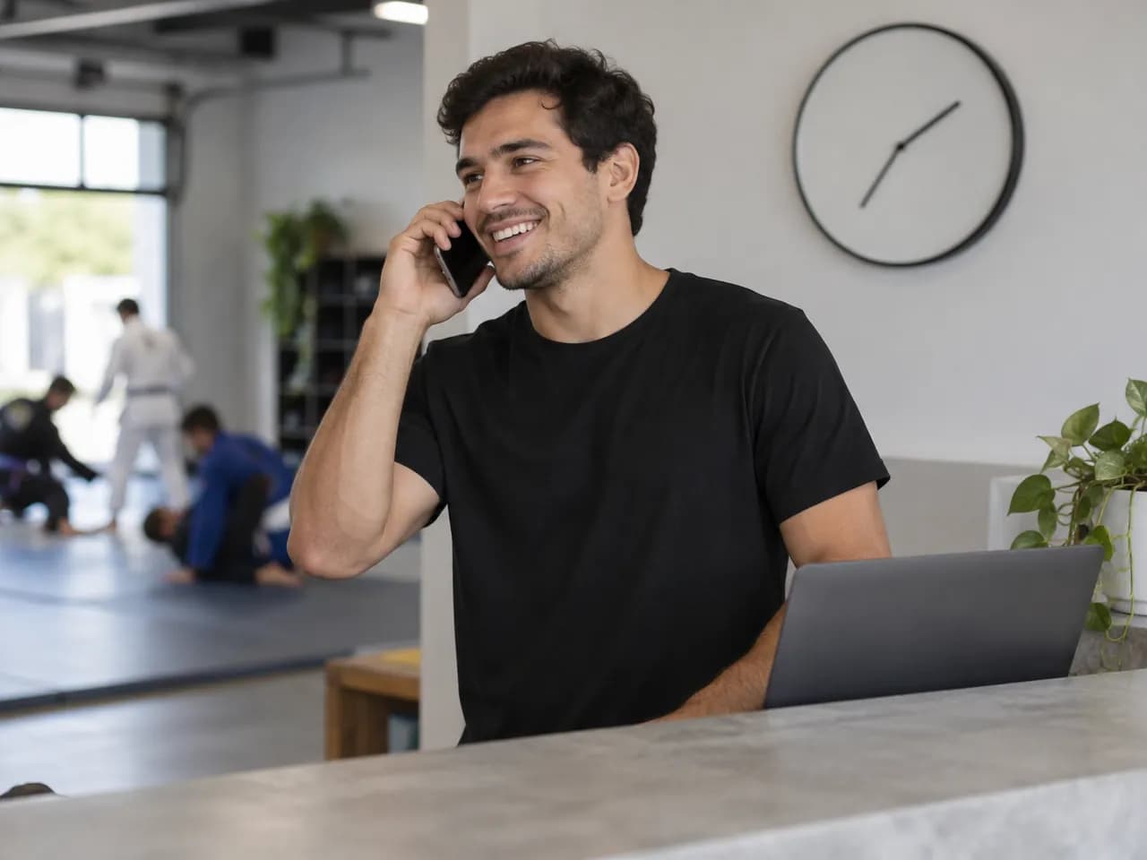A front-desk staff member smiles on a phone call, a wall clock behind him.