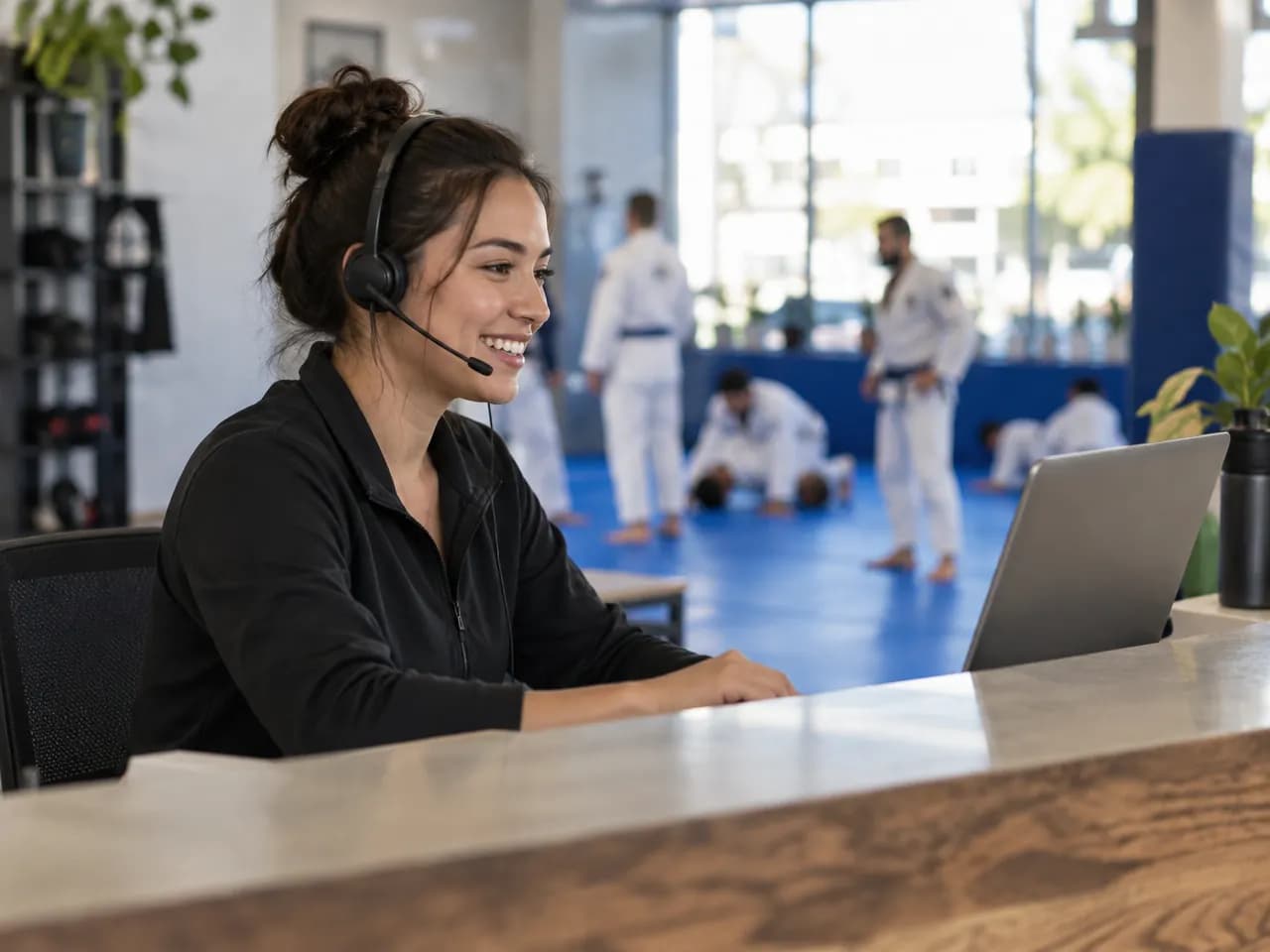 A front-desk staff member in a headset talks and smiles at the reception counter, laptop open, class on the mats behind.