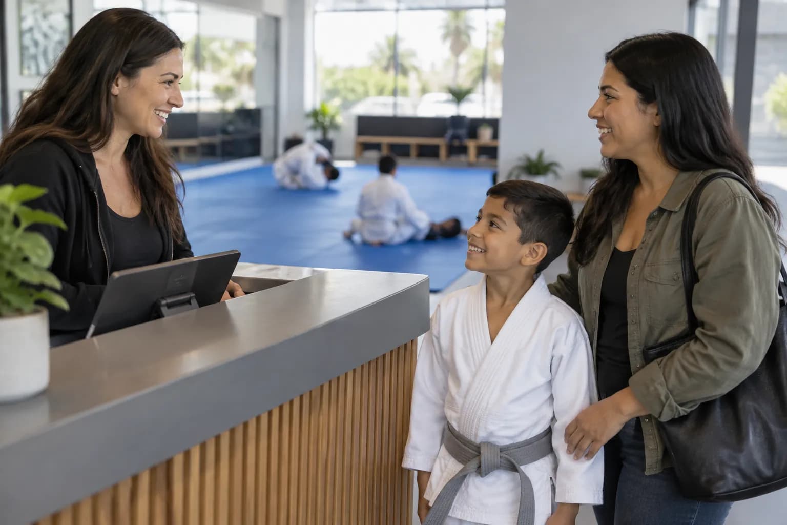 A front-desk staff member greets a mother and her son, who wears a white kids gi with a grey belt.